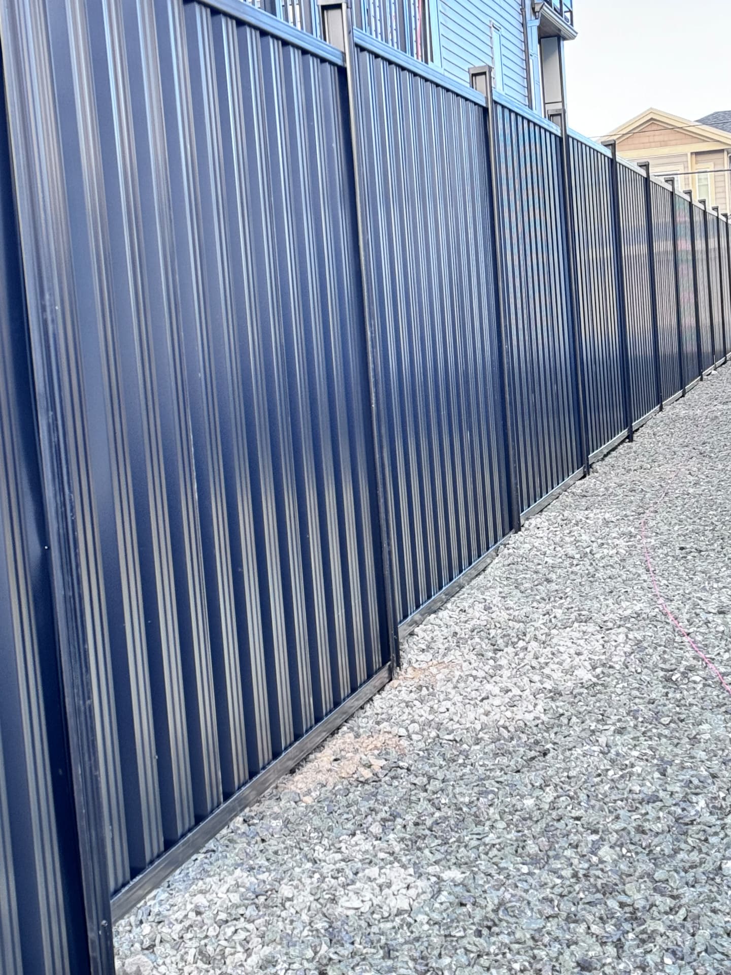 Tall black corrugated metal privacy fence running along a gravel side yard