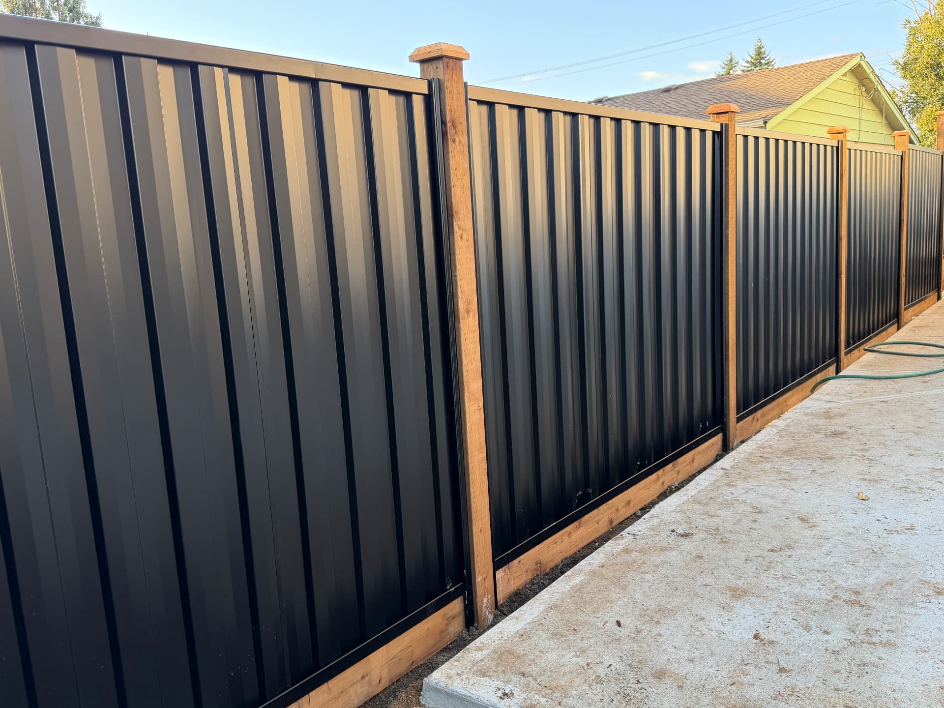 Black corrugated metal privacy fence framed by cedar posts and caps beside a concrete pad