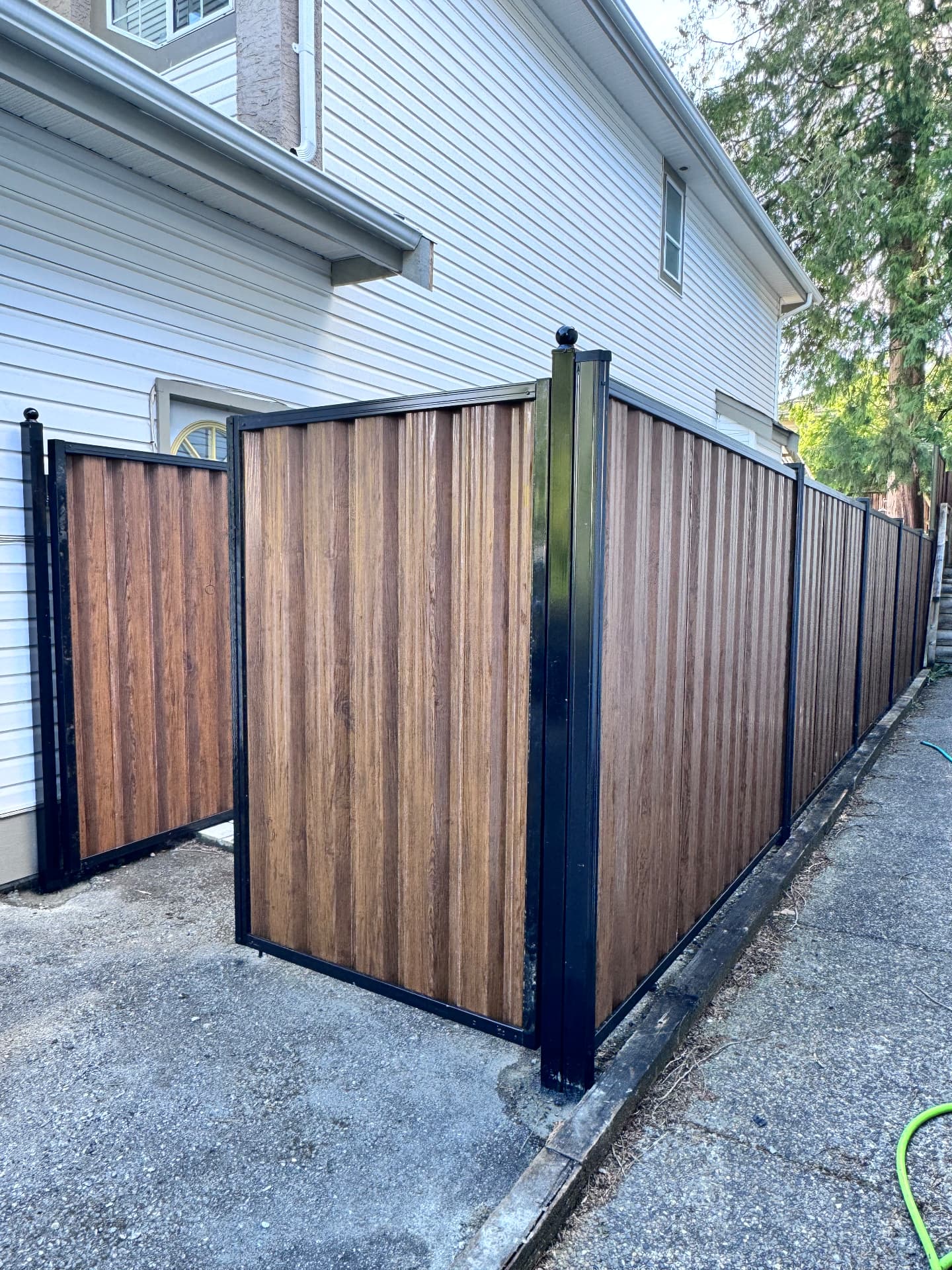 Woodgrain-finish corrugated metal fence and swing gate framed in black aluminum beside a house