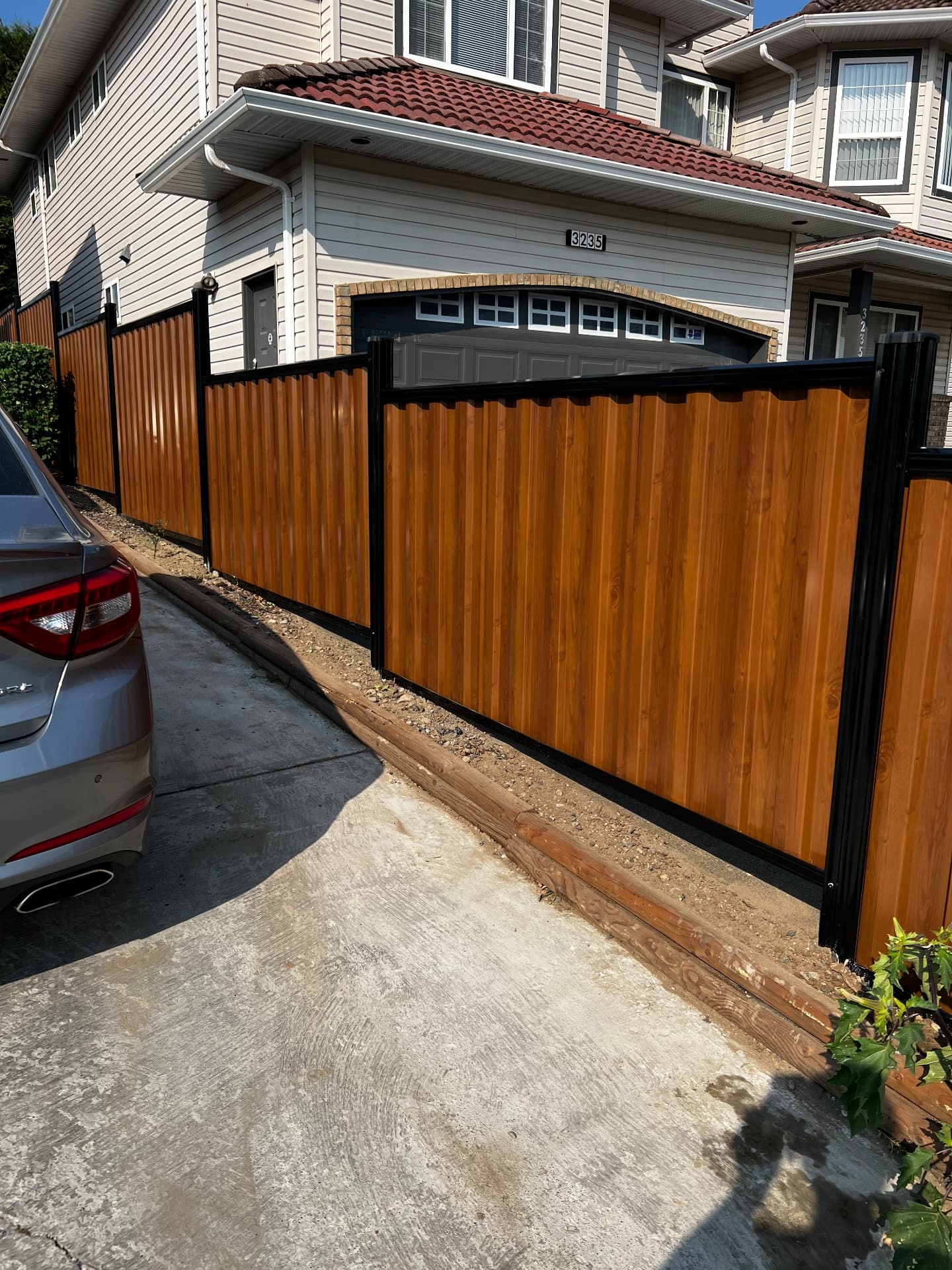 Black-framed woodgrain corrugated metal fence panels lining a residential driveway