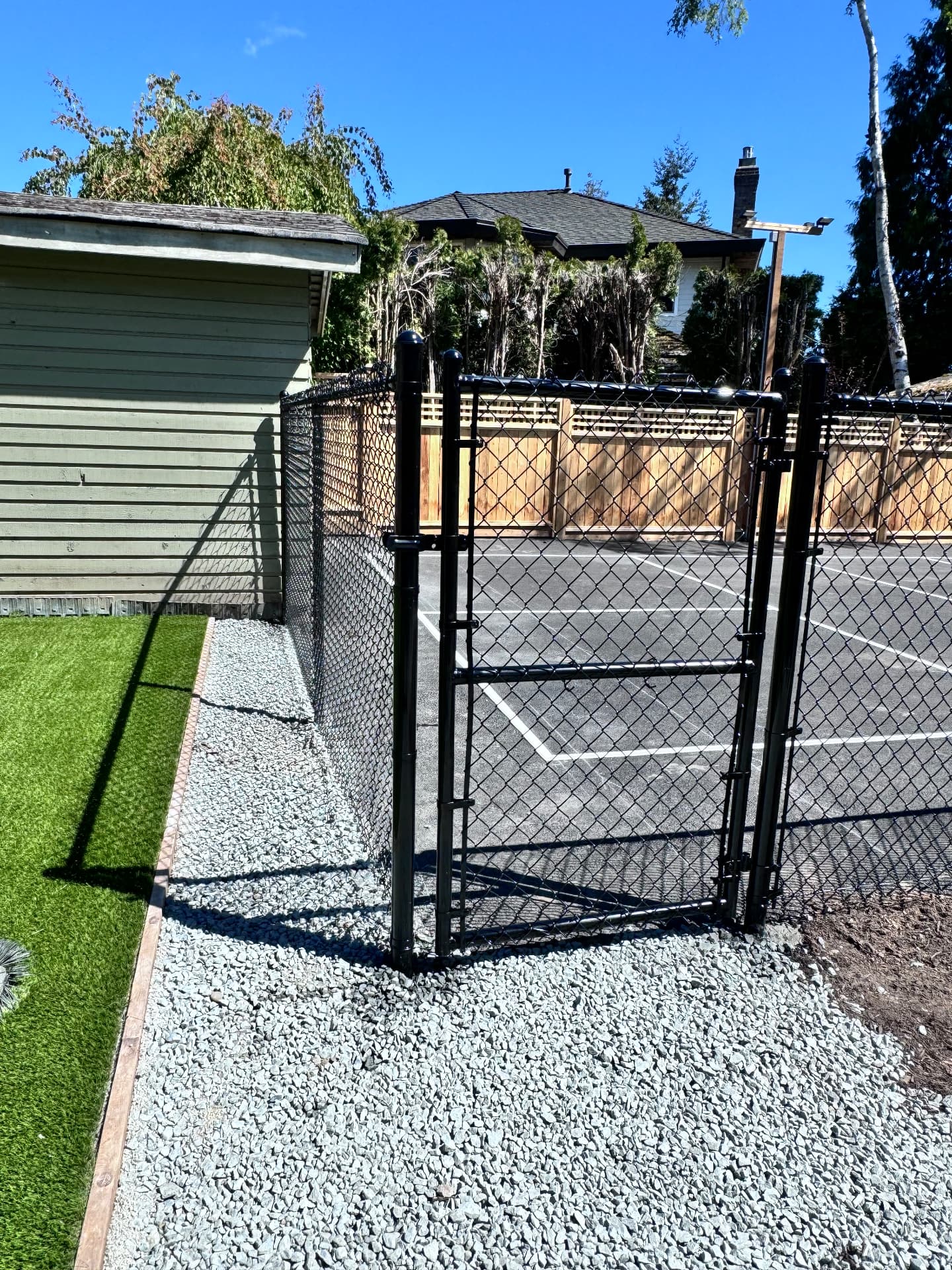 Black vinyl-coated chain link fence and gate set on a gravel bed beside artificial turf